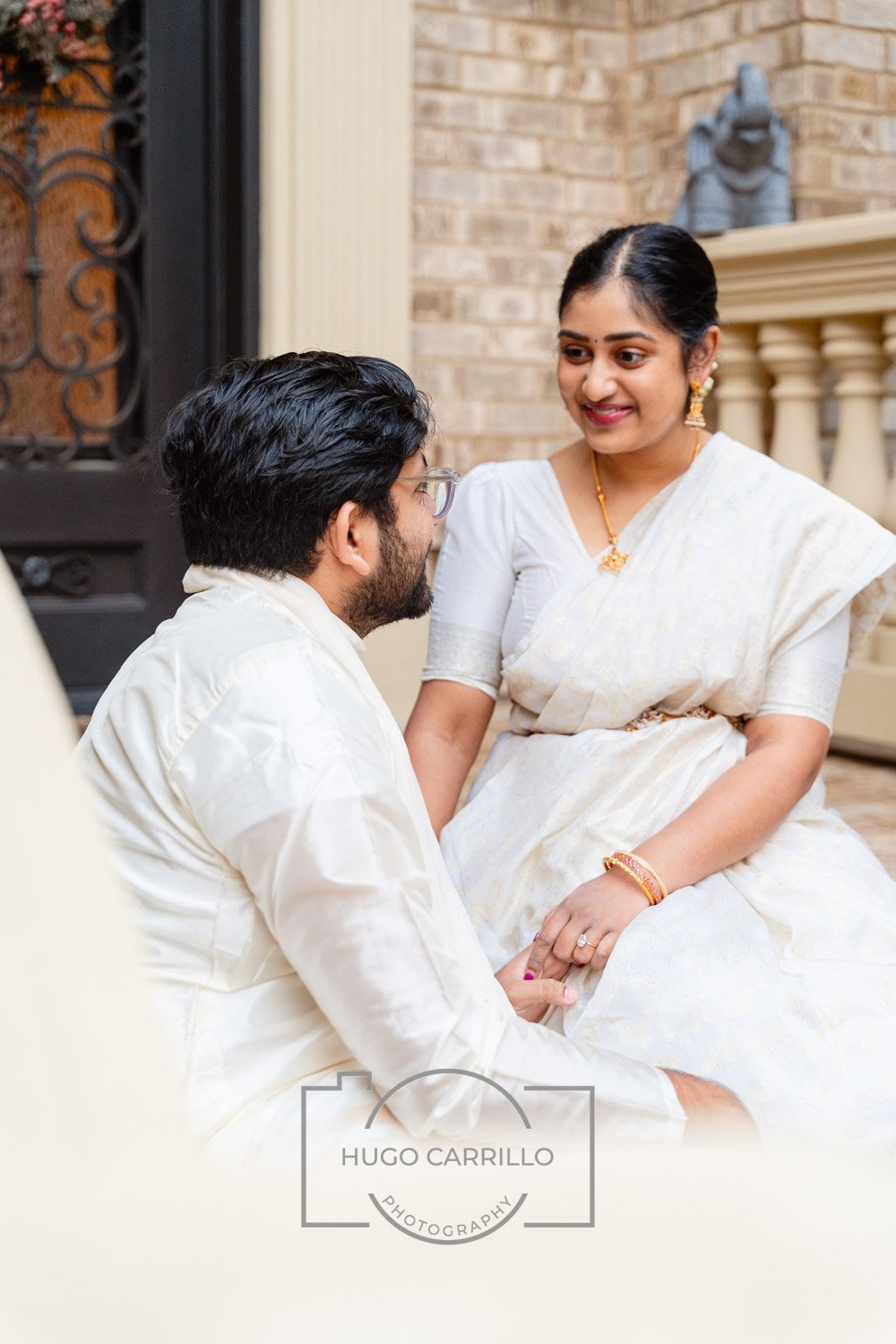 A man and woman dressed in traditional South Asian attire sharing an intimate moment outside near a brick wall and ornate railing. The man is sitting on the ground, looking up at the woman who is sitting on a step, holding hands and smiling at him.
