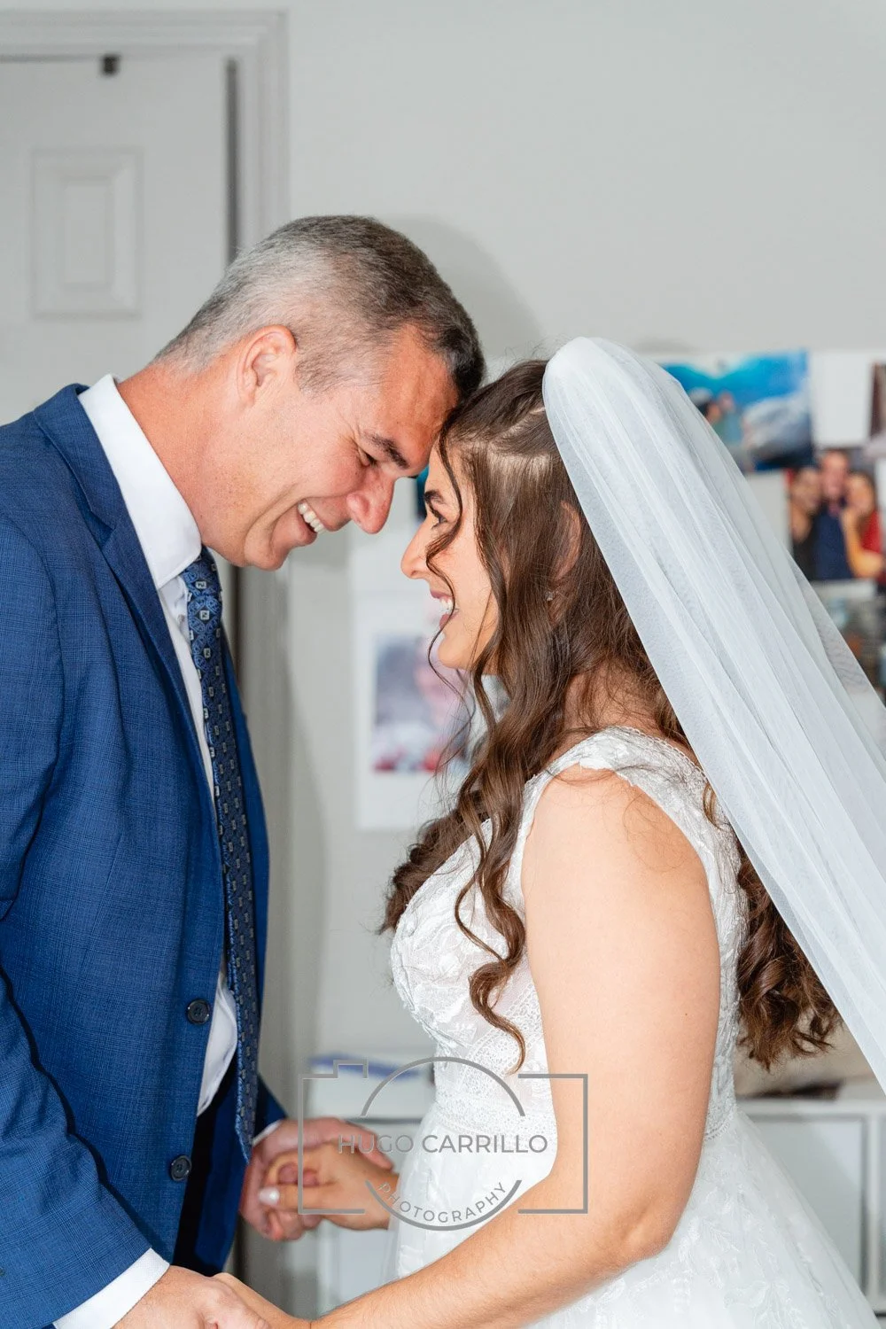 A groom and bride smiling and touching foreheads during their wedding, holding hands inside a room.