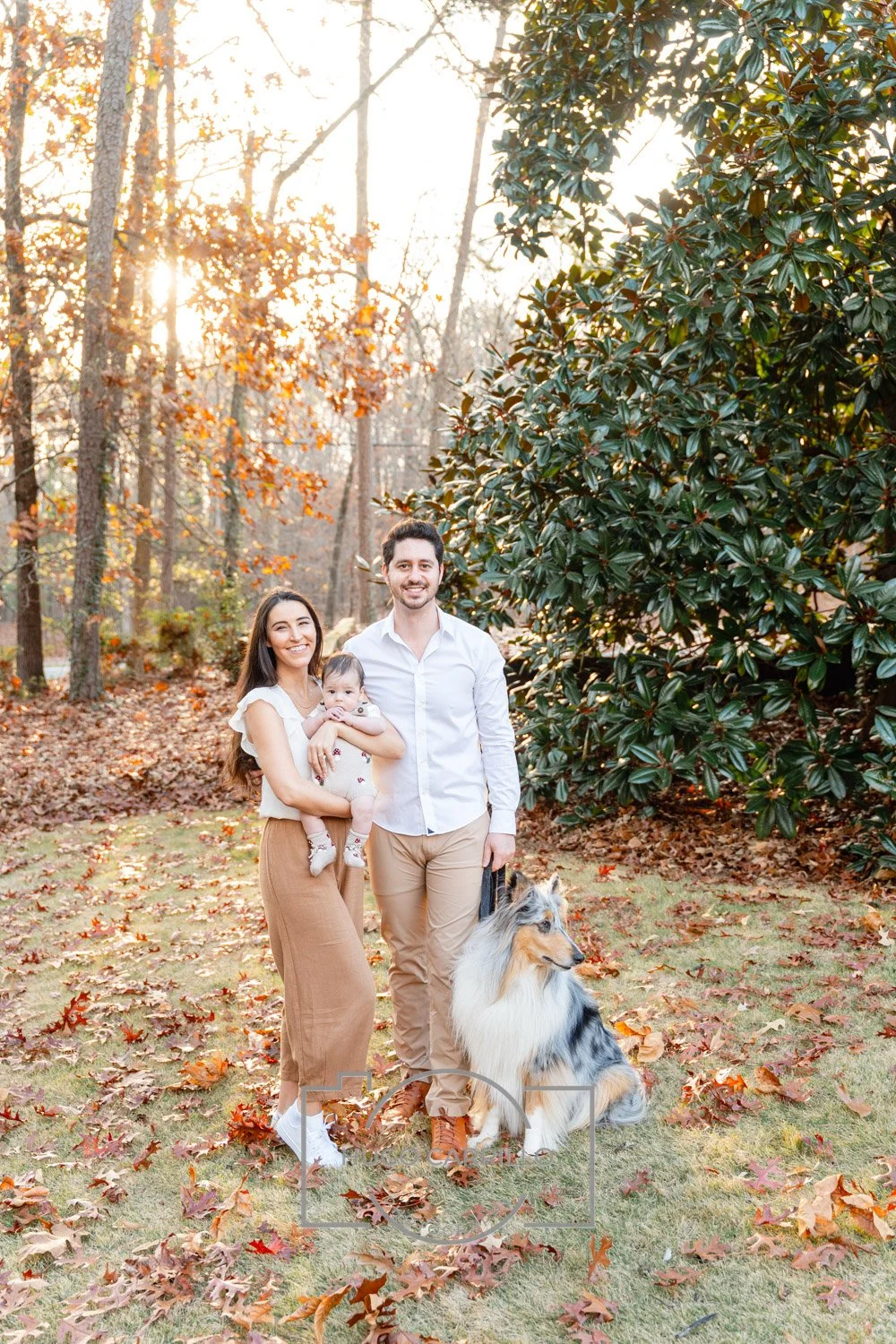 A family of three with a dog, standing outdoors in a wooded area during fall, with sunlight filtering through trees and fallen leaves on the ground.