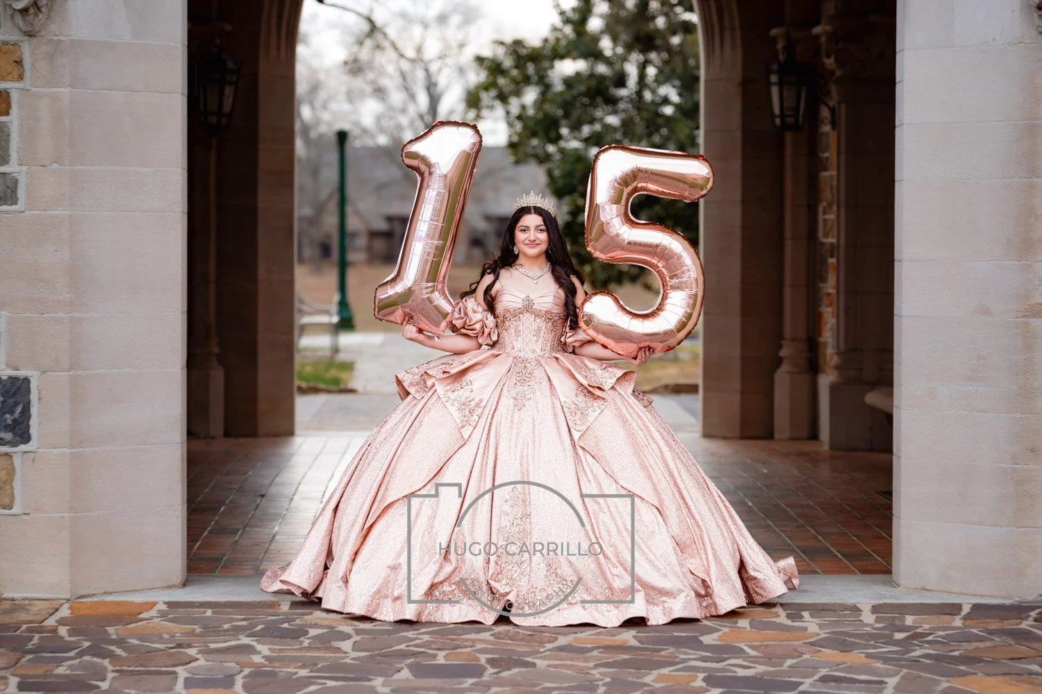 A quinceañera in a pink quinceañera dress holding rose gold balloons shaped as the numbers 1 and 5, standing in a stone archway outdoors.