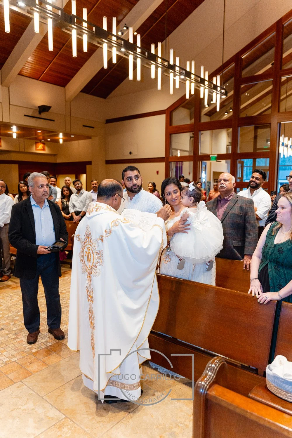 Priest conducting a religious ceremony in a church with a congregation, including a woman holding a young girl dressed in white, and various attendees watching.