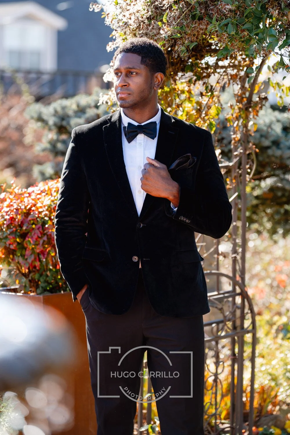 A man dressed in a black velvet tuxedo jacket with a white shirt and black bow tie, standing outdoors among autumn foliage, holding the lapel of his jacket.