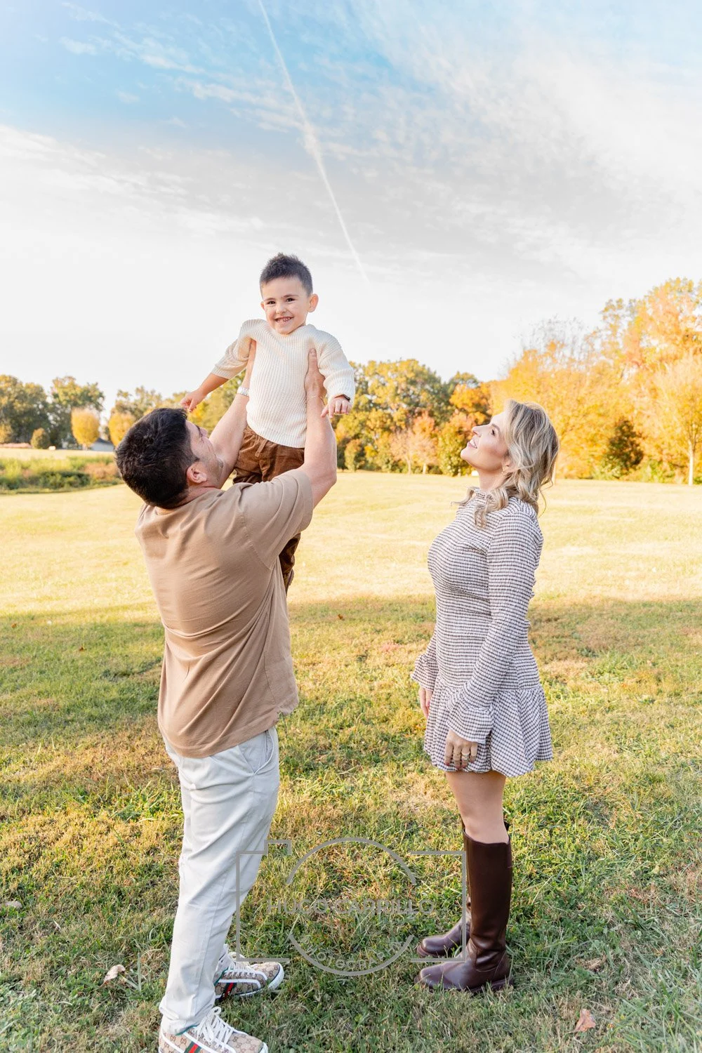 A family of three outdoors in a sunny field with autumn trees in the background. A man lifts a smiling young boy wearing a cream sweater and brown pants, while a woman in a checkered dress and tall boots looks on.