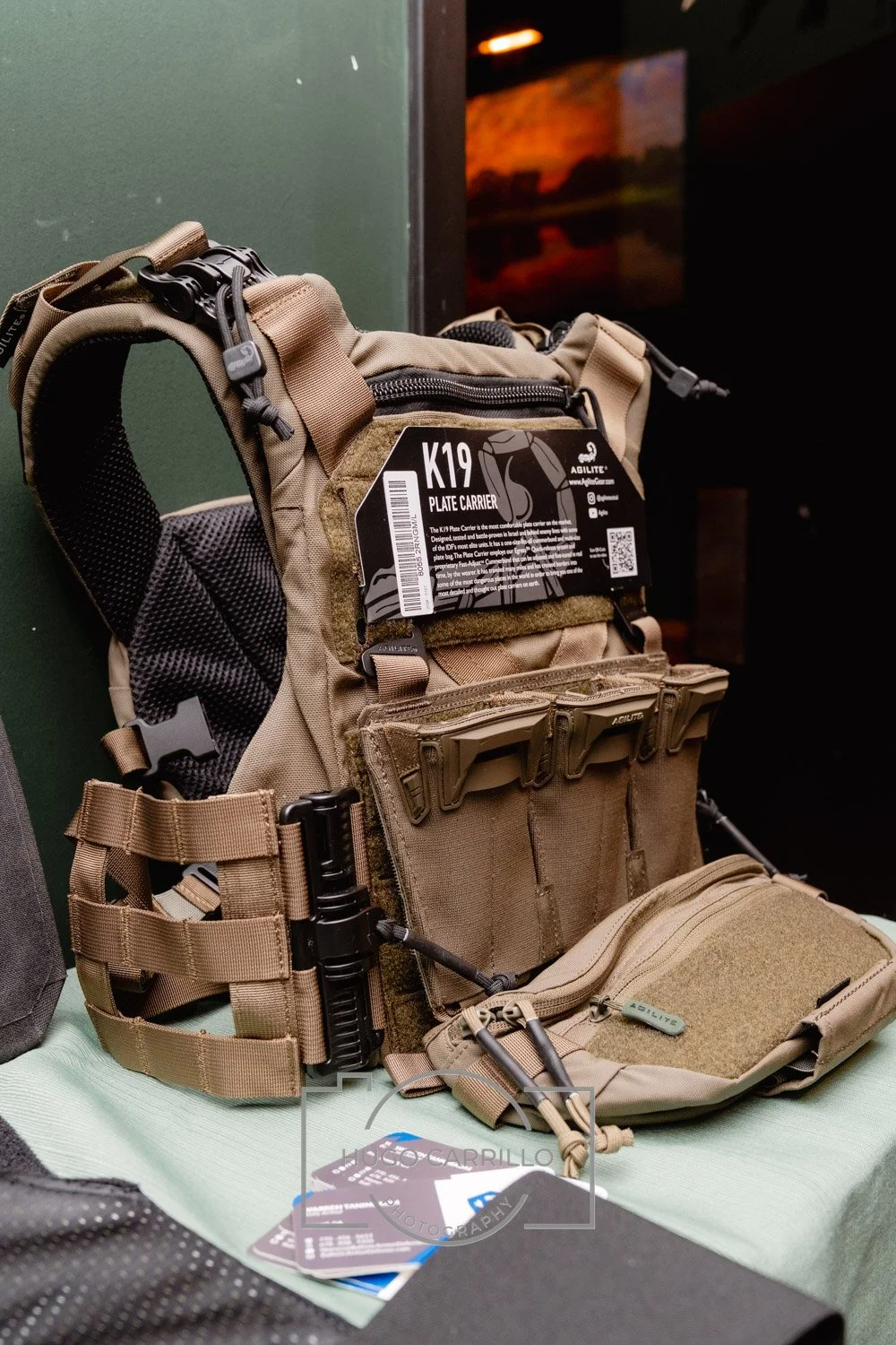 Tactical beige plate carrier vest with multiple pouches and straps, labeled 'K19 Plate Carrier' by Agilite, displayed on a table with informational tags.