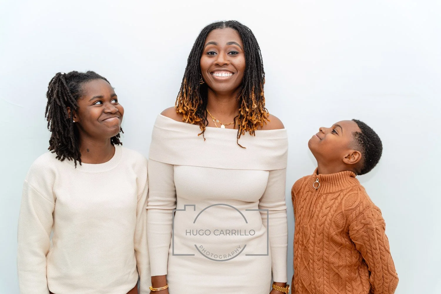 A woman with shoulder-length, curly hair wearing an off-the-shoulder cream dress smiling at two children, a girl with dreadlocks in a cream sweater and a boy in a brown sweater looking up at her, all standing against a plain white wall.
