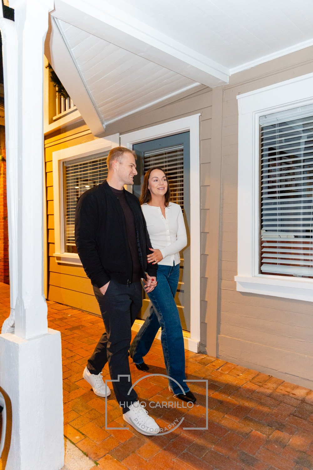 A couple walking outside a house at night, smiling and holding hands, on a brick walkway near a beige house with white trim and windows with blinds.