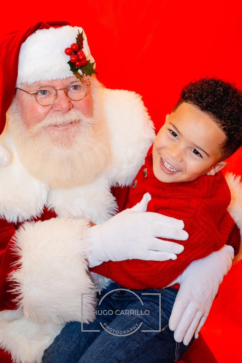 A young boy and Santa Claus are smiling happily during a Christmas celebration, with Santa wearing his traditional red suit and hat decorated with holly berries. The boy is dressed in a red sweater and jeans, sitting on Santa's lap against a red back