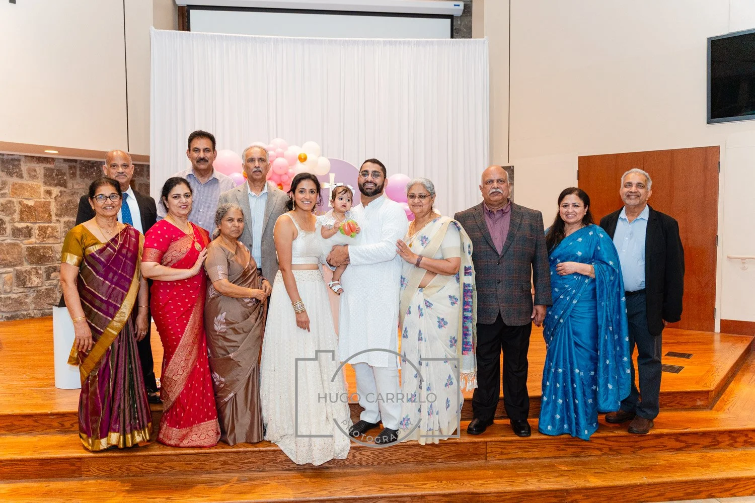 A group of people dressed in traditional Indian attire standing on a wooden stage, celebrating a special occasion with pink, white, and gold balloons and a white draped backdrop.