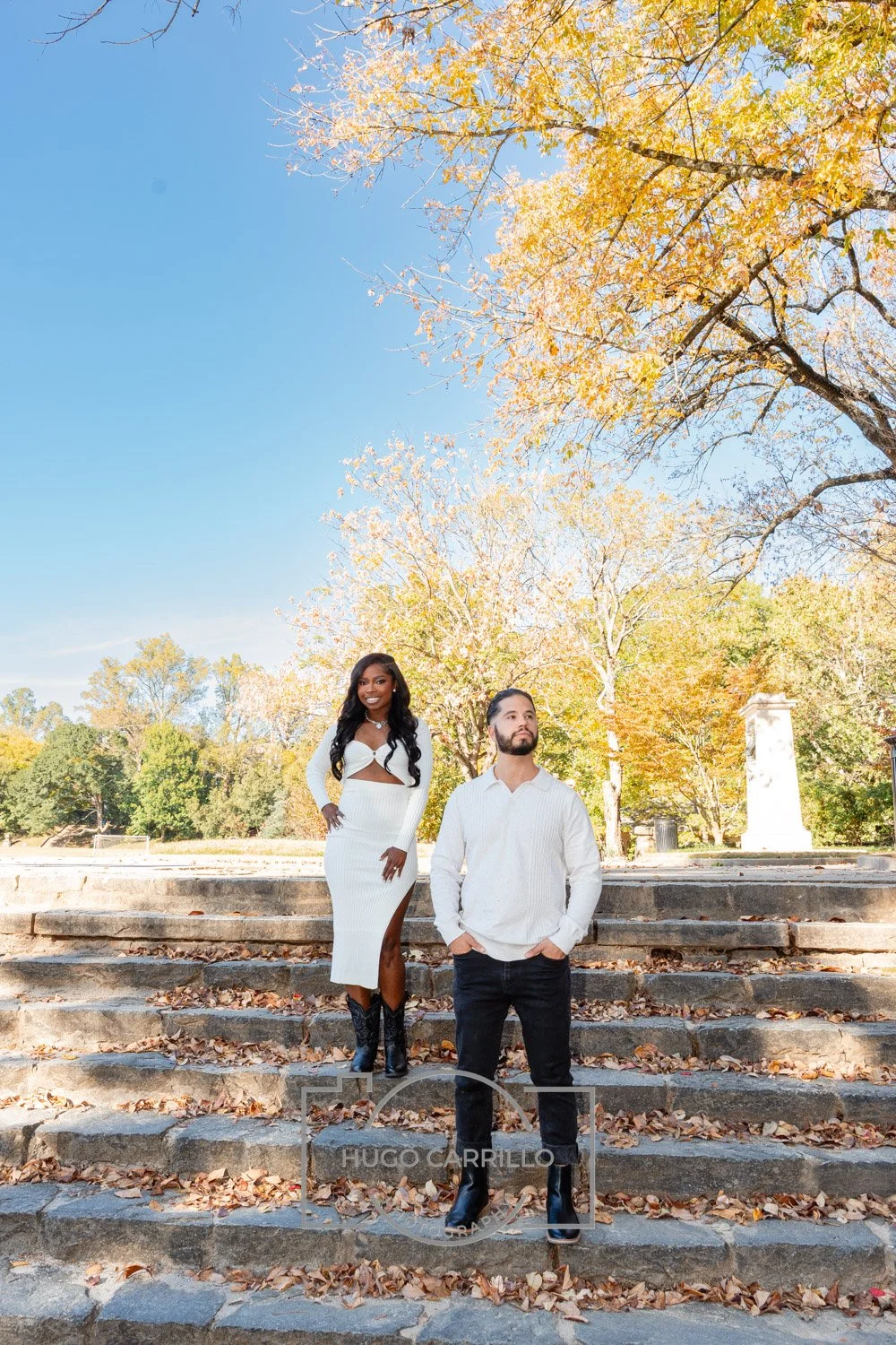 A woman and a man standing on stone stairs in a park during autumn, with colorful fall leaves on trees and a clear blue sky in the background.
