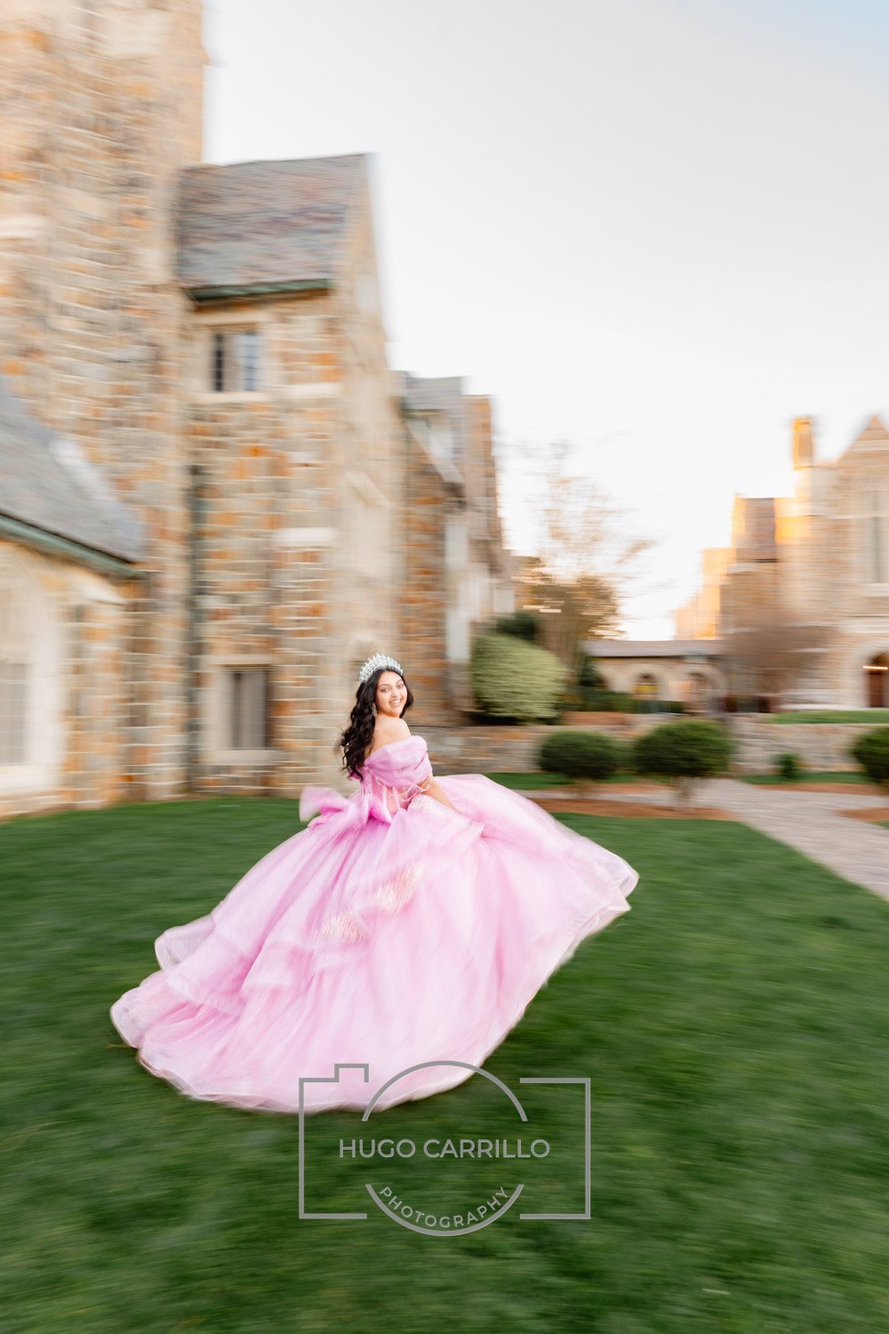 A young woman in a pink quinceañera dress and tiara spinning on a lawn in front of a stone building.