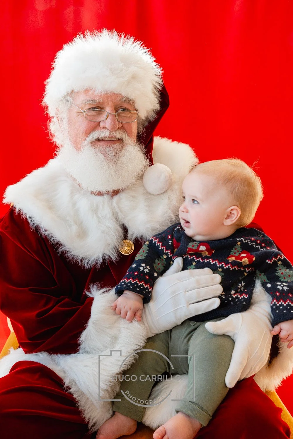 Santa Claus in a red velvet suit with white fur trim holding a young child with blonde hair and a Christmas sweater against a red backdrop.