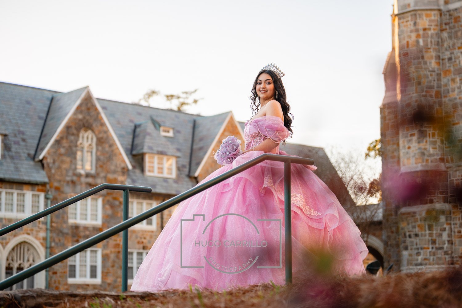 Young woman in a pink princess-style gown and tiara standing on a staircase outside a stone castle-like building, holding a bouquet of pink roses and smiling.
