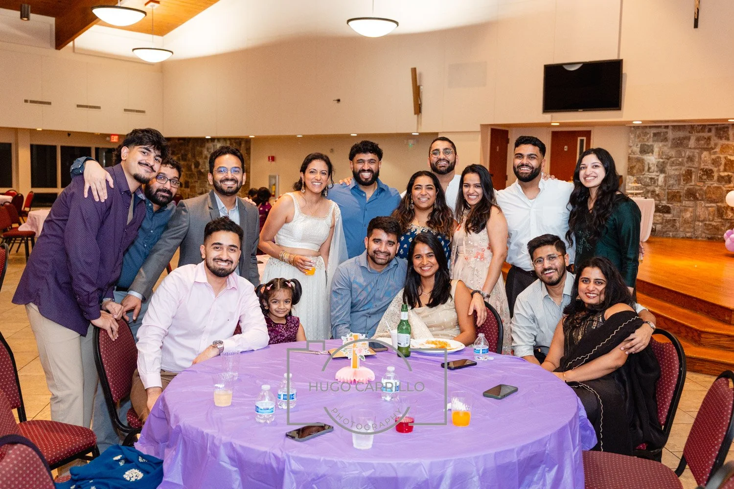 A group of 15 people, including men, women, and a young girl, smiling and posing around a table with a purple cloth at a celebration or party in a banquet hall, with some holding drinks.