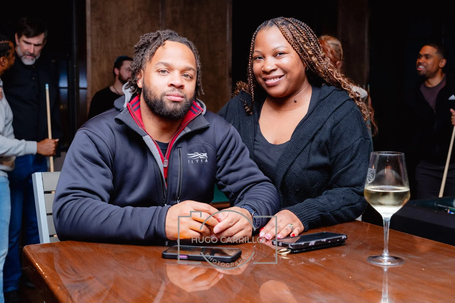 A man and woman sitting at a wooden table, the man in a dark jacket with a logo, and the woman smiling in a black sweater, with a glass of wine and two smartphones on the table, with other people in the background.
