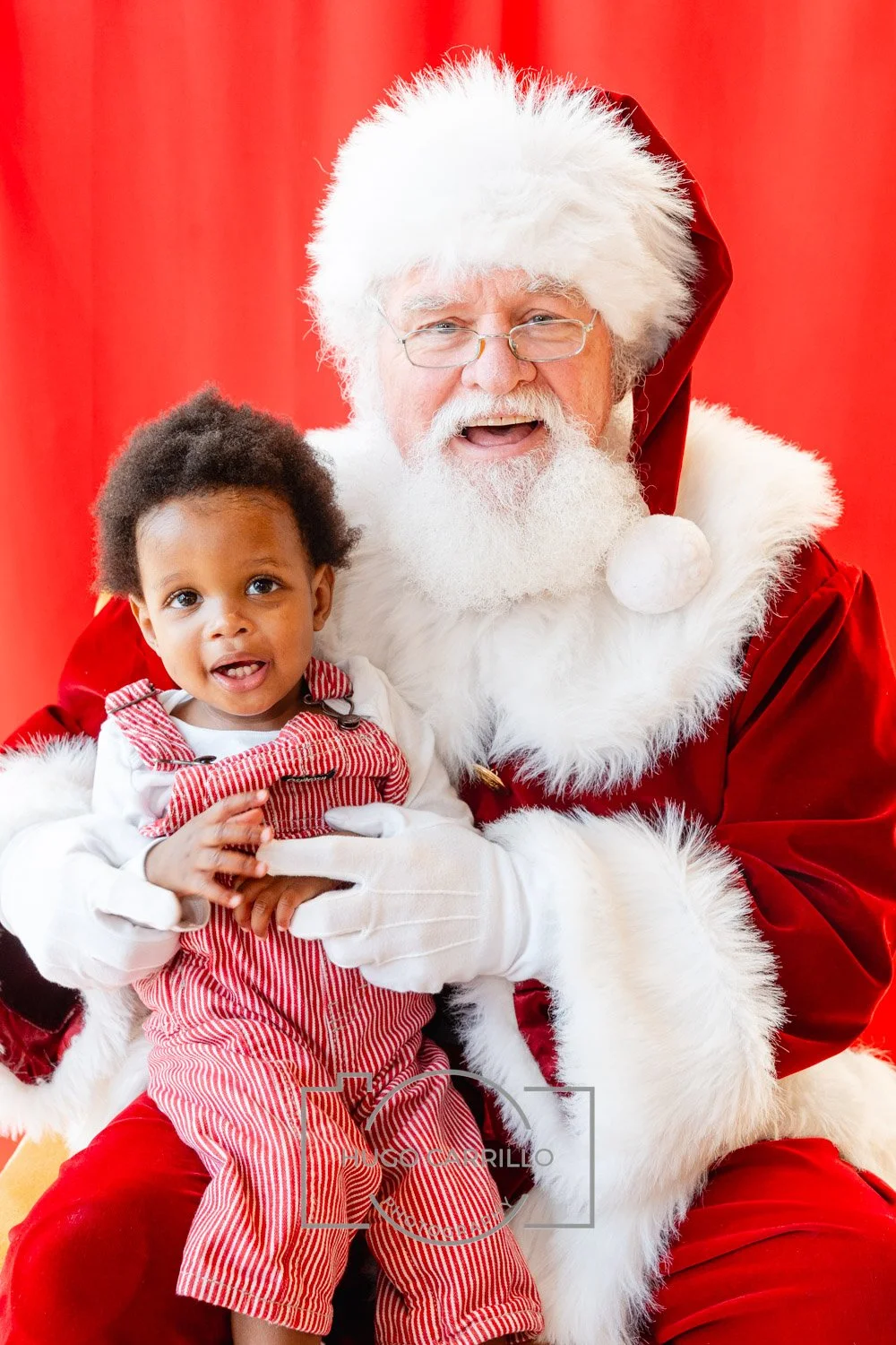 A young child sitting on Santa Claus's lap during a Christmas event, with Santa dressed in his signature red and white suit, hat, and glasses, against a red background.