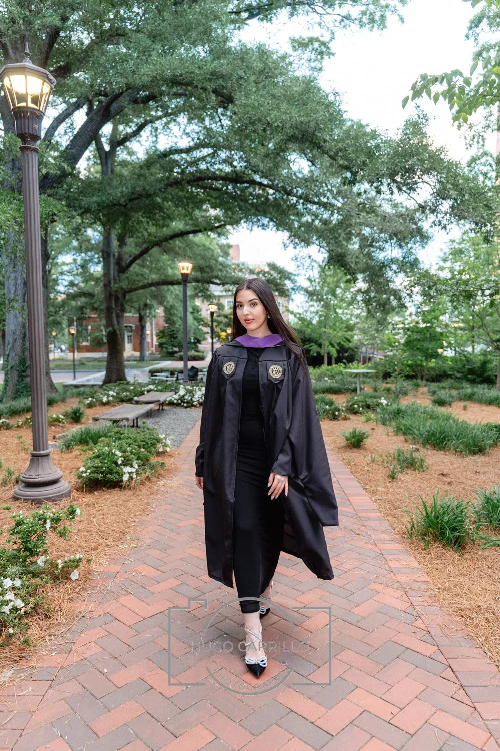 Young woman in graduation gown and cap walking on brick pathway in a park with trees and lampposts at dusk.