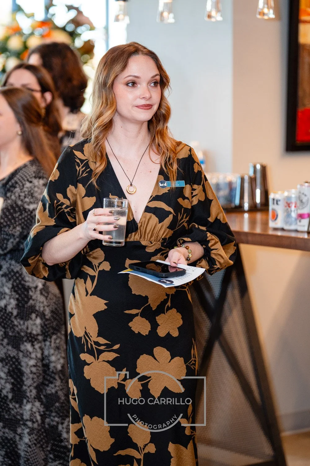 A woman with wavy, shoulder-length hair wearing a black dress with large tan floral patterns, holding a glass of water and some papers and a phone, at a social event or gathering.