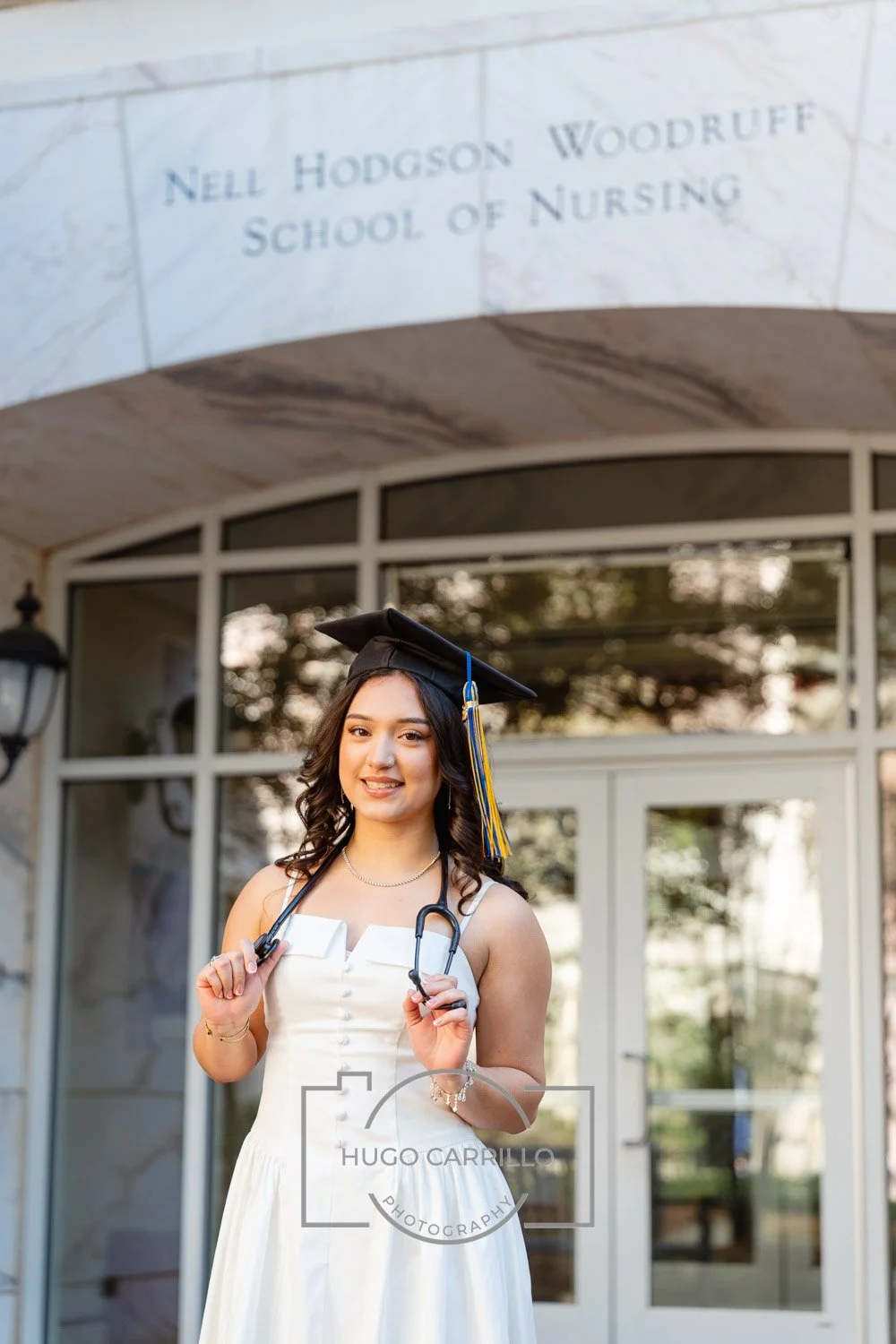 Young woman in a white dress and graduation cap holding a stethoscope, standing outside a building with the sign 'Nell Hodgson Woodruff School of Nursing' above the entrance.