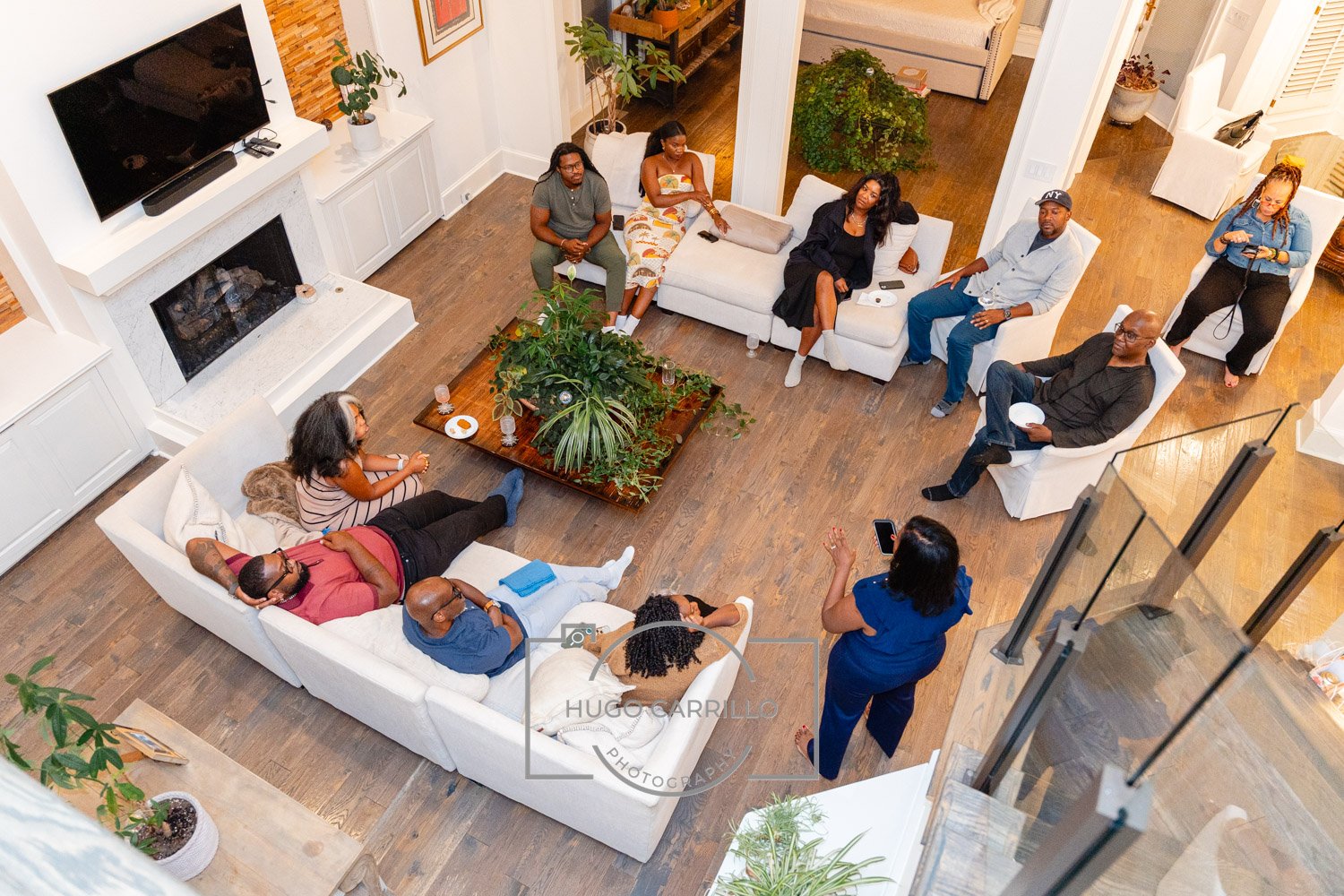 People gathered in a living room for a meeting, some sitting on sofas, others standing or seated on the floor, with a coffee table, plants, and a fireplace in the background.