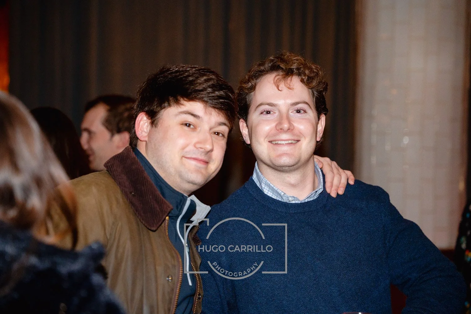 Two young men smiling at an event, one with dark hair wearing a leather jacket and the other with curly hair in a blue sweater, standing close together with one arm around each other's shoulders.