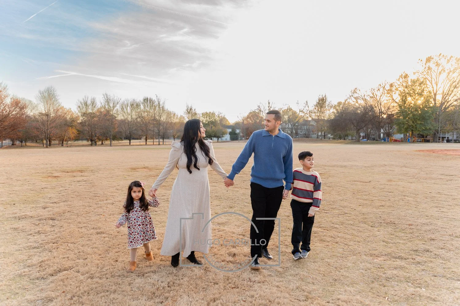 A family of four walking hand in hand in a park during sunset. The mother, father, and two children are smiling and holding hands.