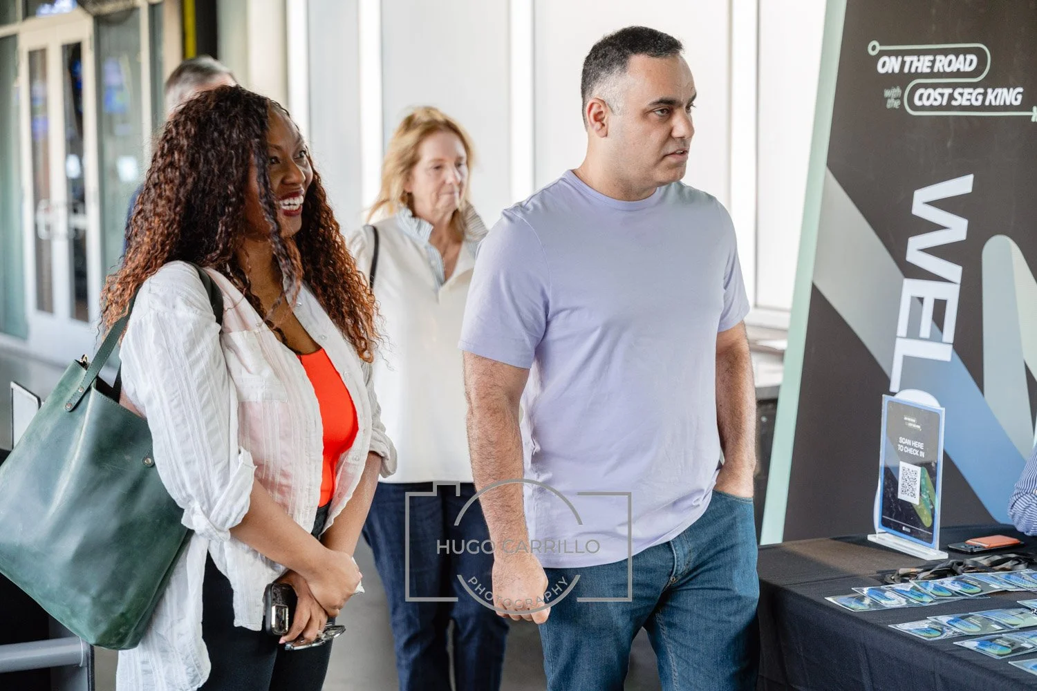 People standing in line at a registration or check-in booth, with a man looking at a display board and a woman smiling.