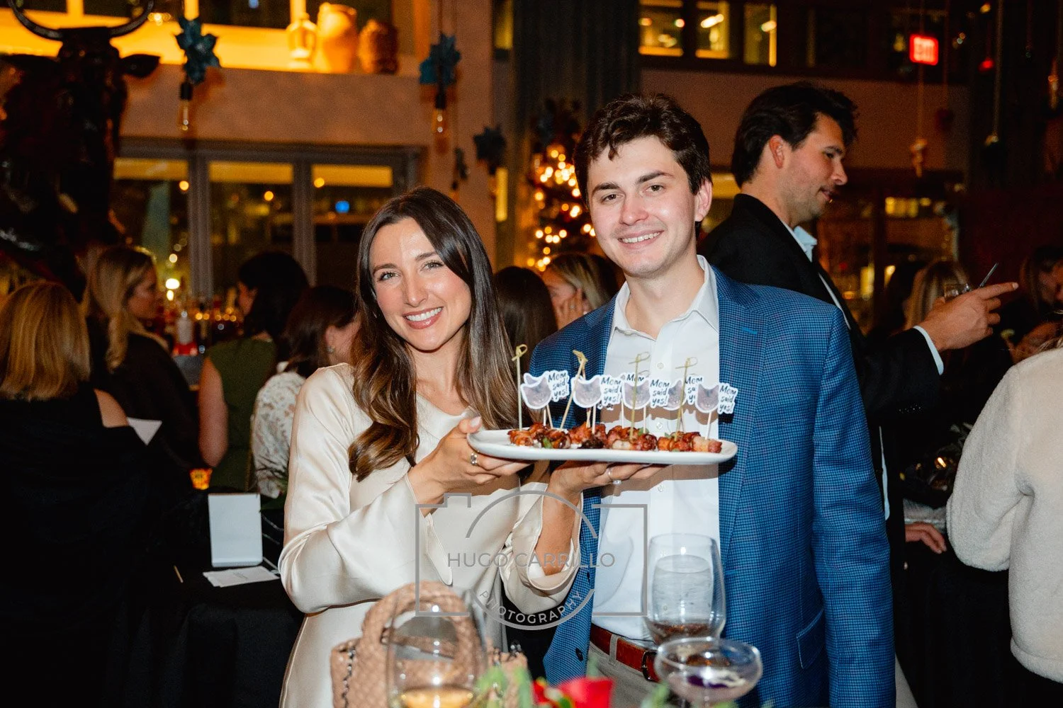 A woman and a man smiling at a celebration, holding a plate of appetizers with small flags that read 'Mom said yes' and 'Dad said yes'.