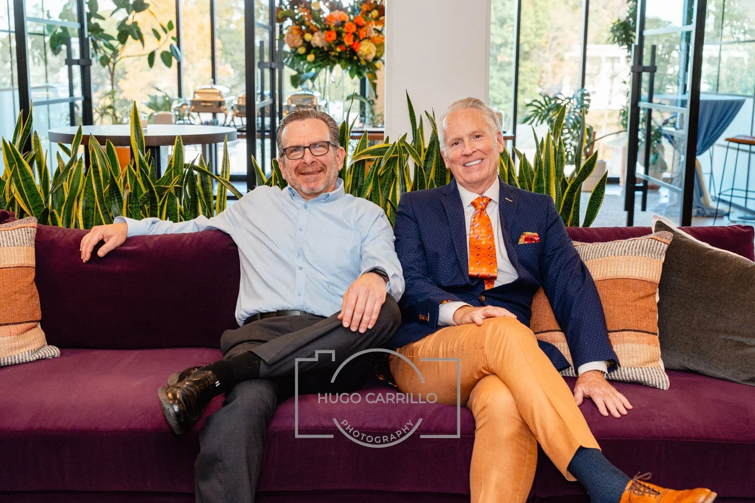 Two men sitting on a purple sofa with yellow and striped throw pillows, smiling, in a modern indoor space with plants and large windows in the background.