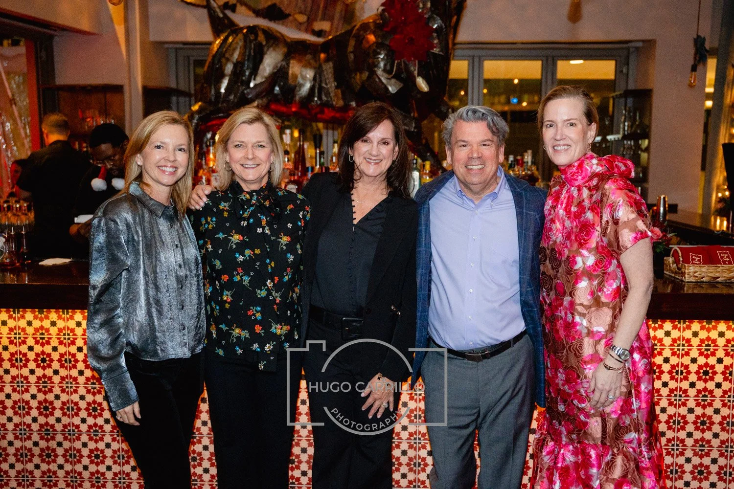 Group of five people standing together at a social event, smiling, in an indoor venue with a bar in the background.