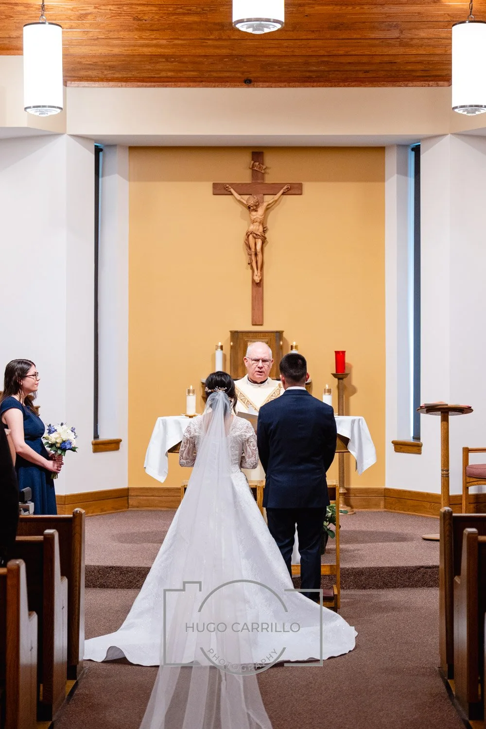 A wedding ceremony taking place inside a church, with a bride in a white wedding gown and veil, a groom in a dark suit, a priest officiating, and a bridesmaid holding a bouquet, all standing at the altar in front of a crucifix.