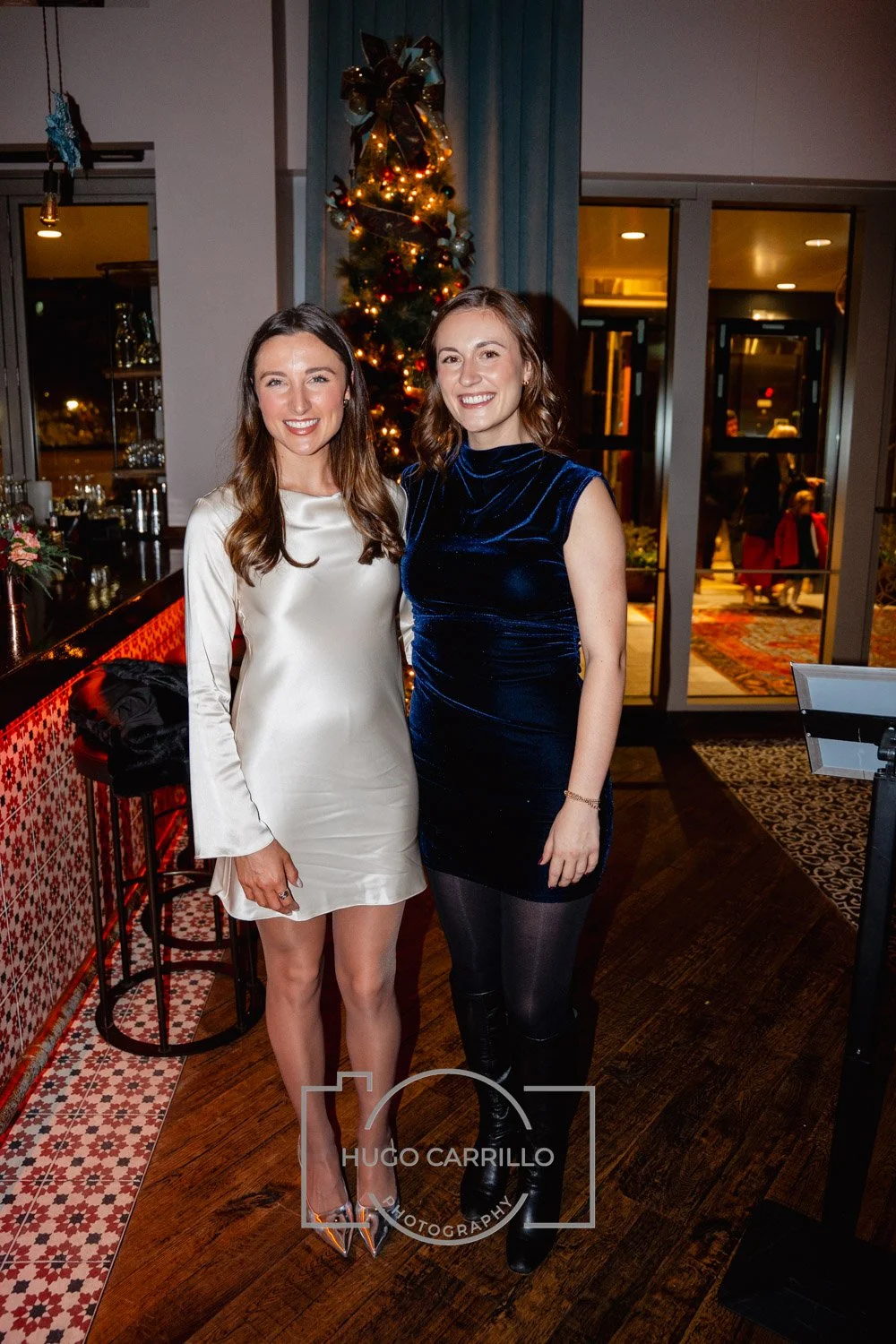 Two women standing inside a restaurant at night, smiling for the camera, with a decorated Christmas tree in the background.