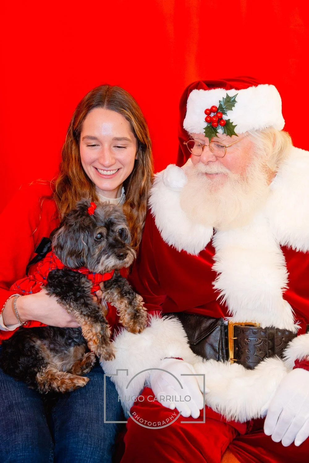 A woman with long red hair and a pet dog sitting beside a man dressed as Santa Claus against a red background.