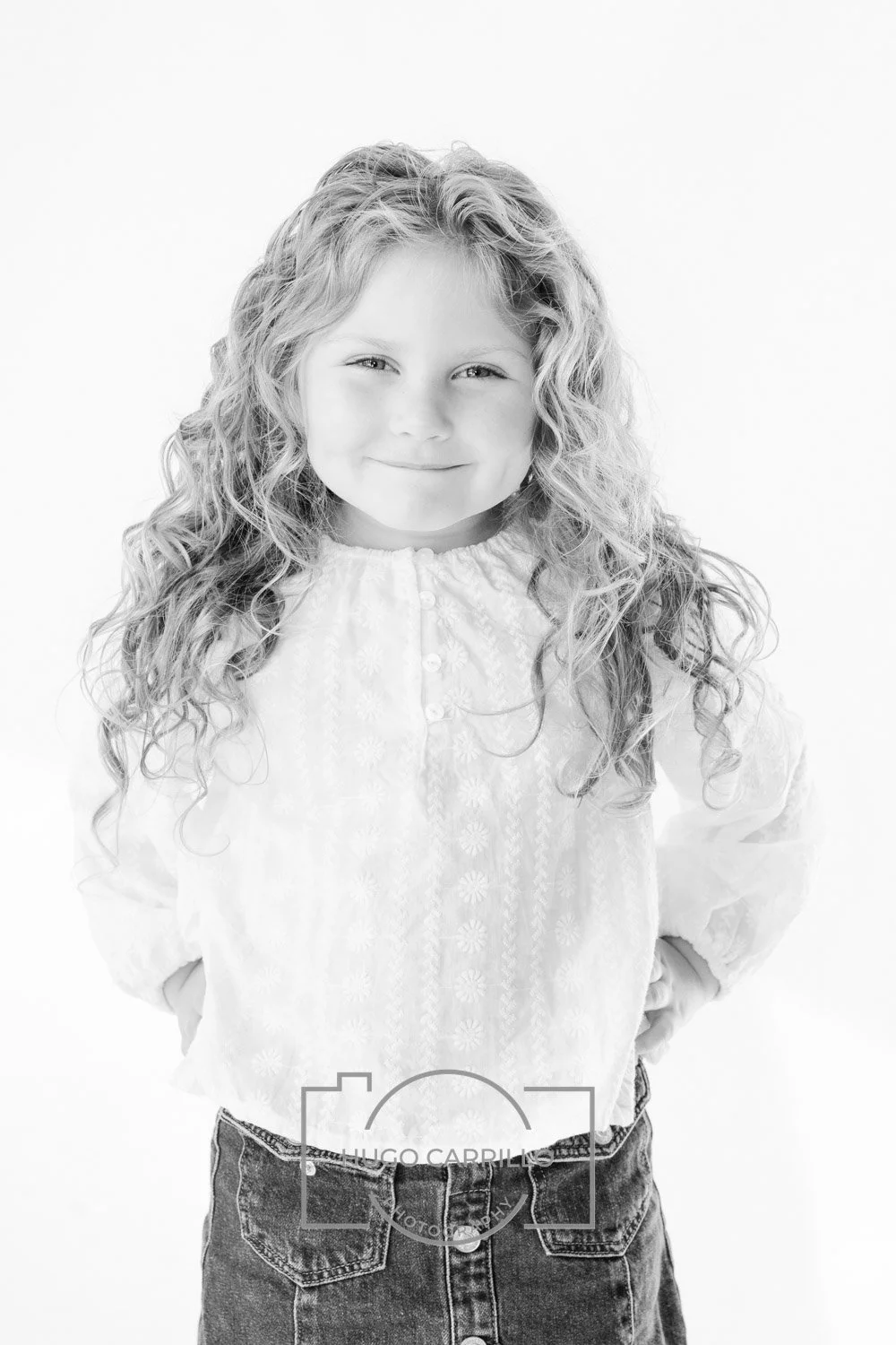 Black and white photo of a young girl with curly hair, smiling, wearing a long-sleeved blouse and denim skirt, with hands in pockets, against a plain background.