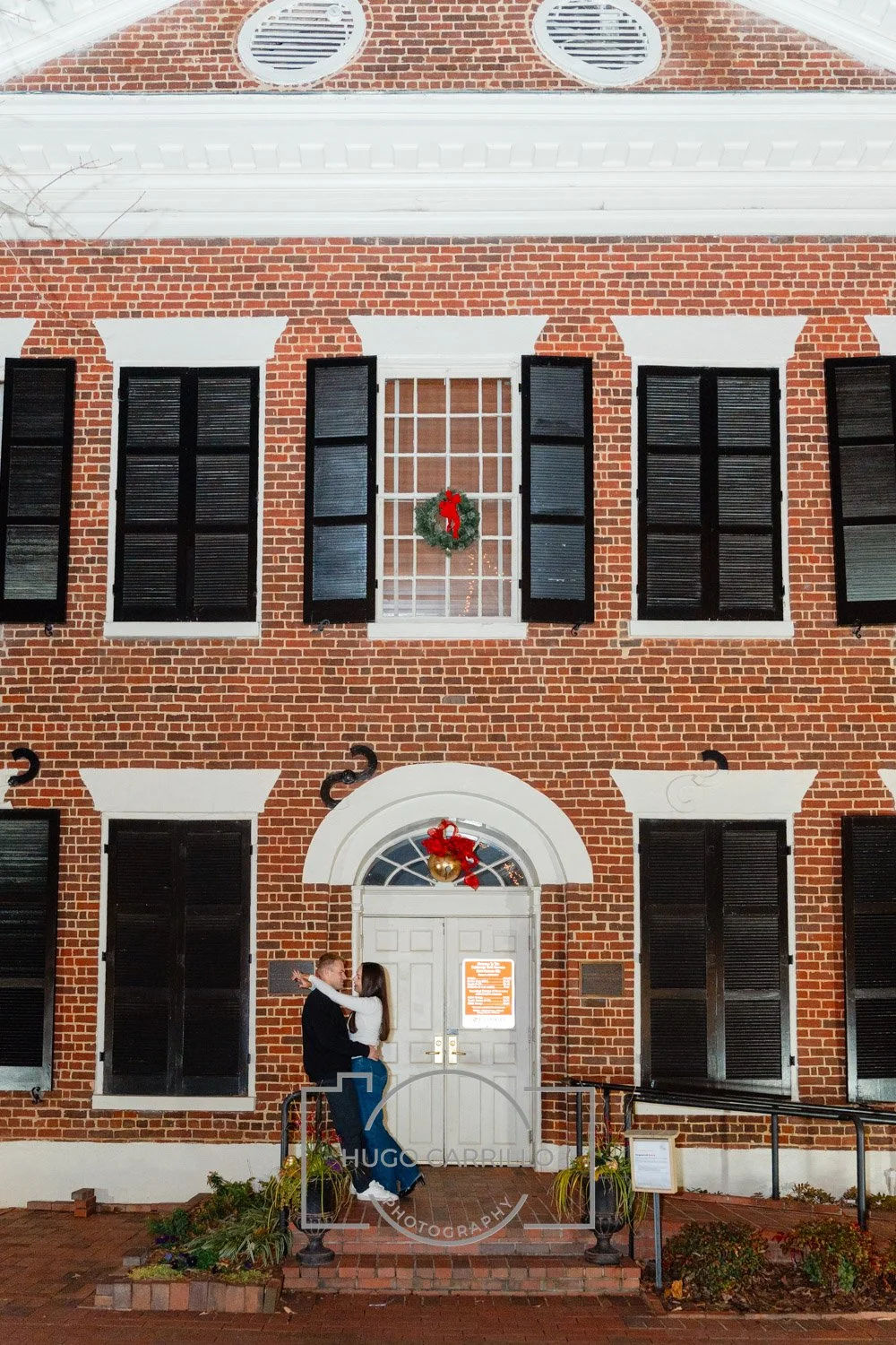 A couple stands in front of a brick building decorated with Christmas wreaths, engaged in a romantic dance or embrace.