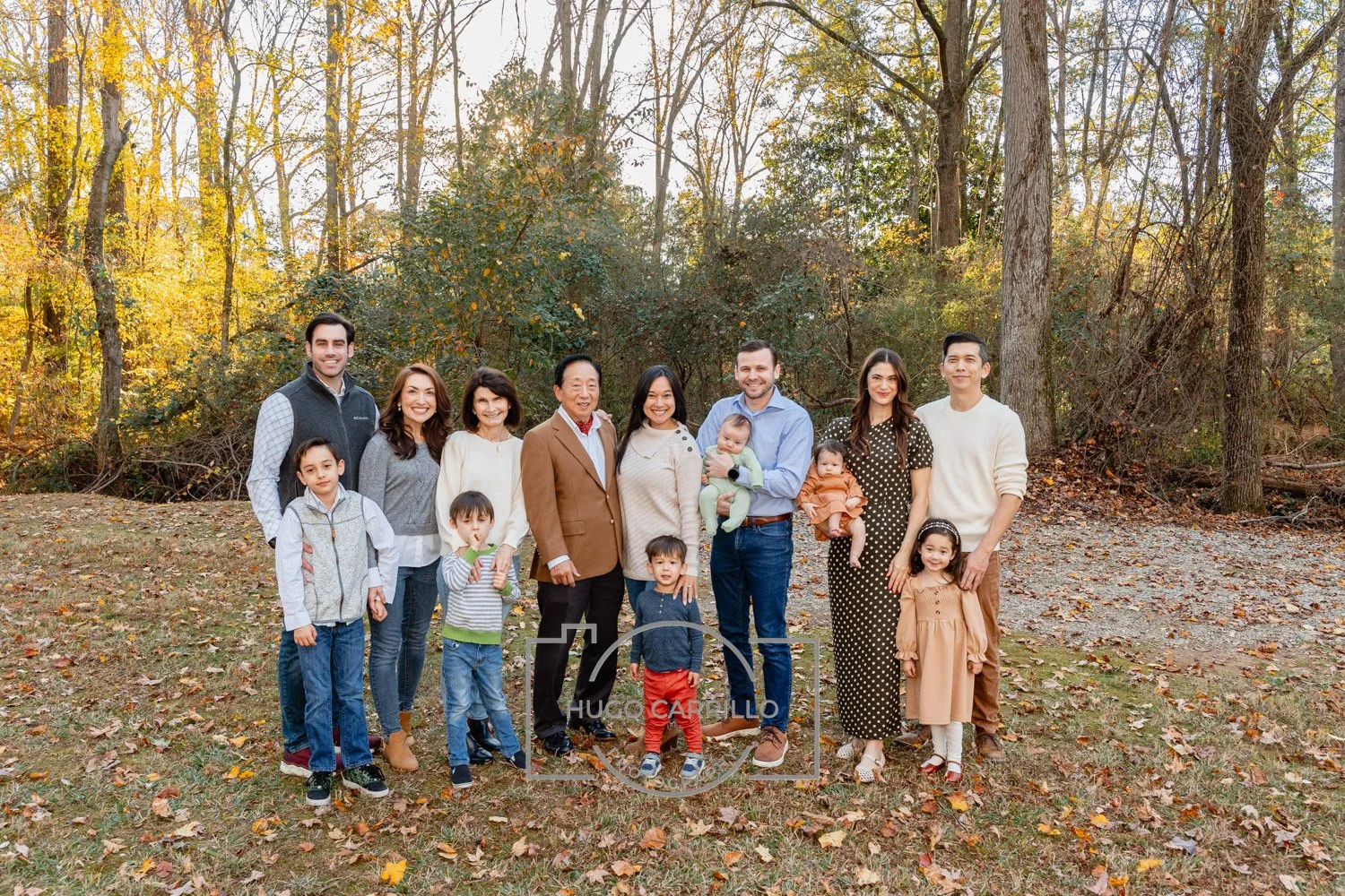 A large multigenerational family standing outdoors in a wooded area during fall, smiling for a group photo.