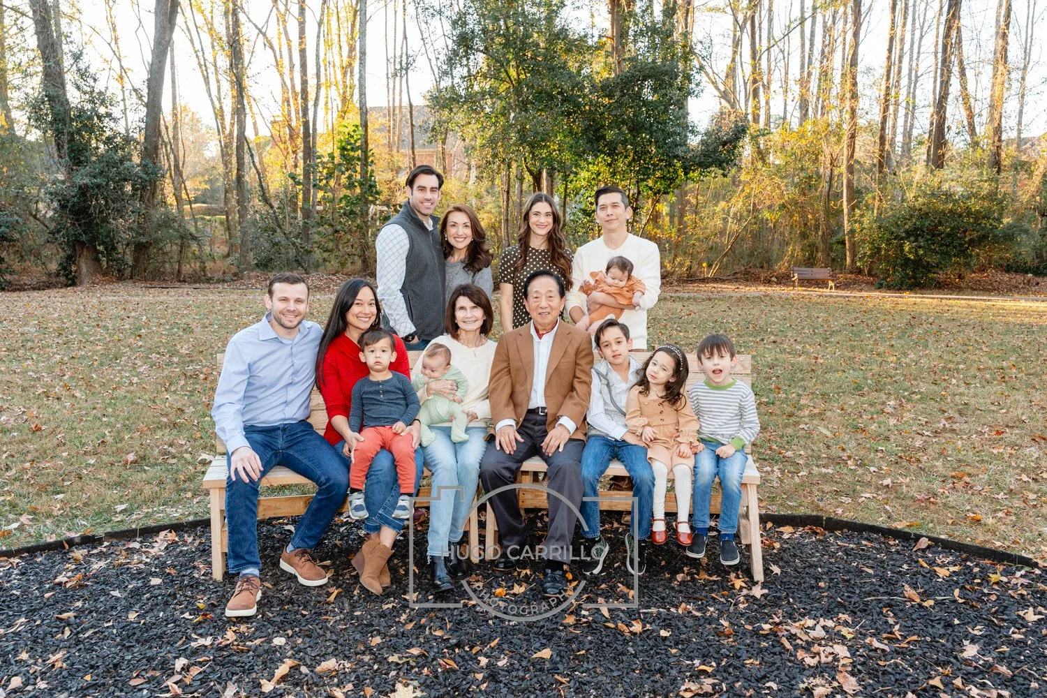 Family group photo outdoors in park during autumn, with adults and children sitting and standing on a wooden bench, surrounded by trees with fall foliage.