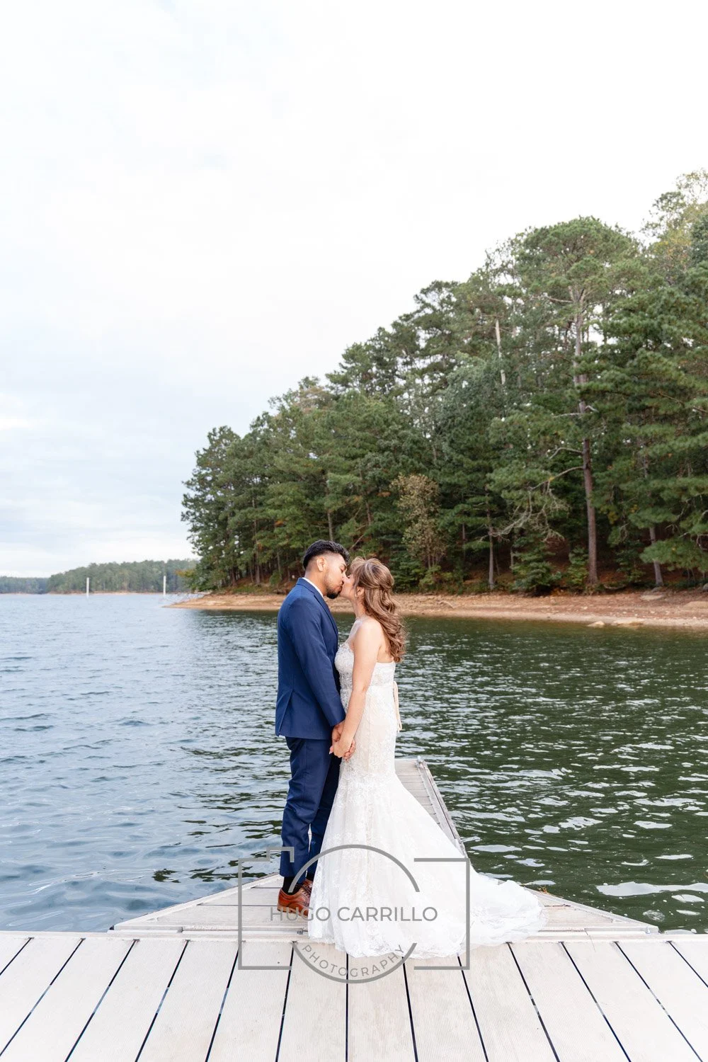 A bride and groom standing on a dock by a lake, kissing, surrounded by trees.