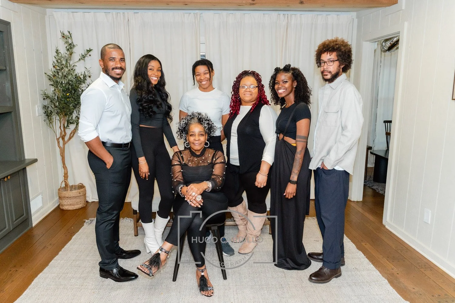 A diverse group of eight people, seven standing and one sitting in front of white curtains in a room with wooden floors, smiling for a photo.
