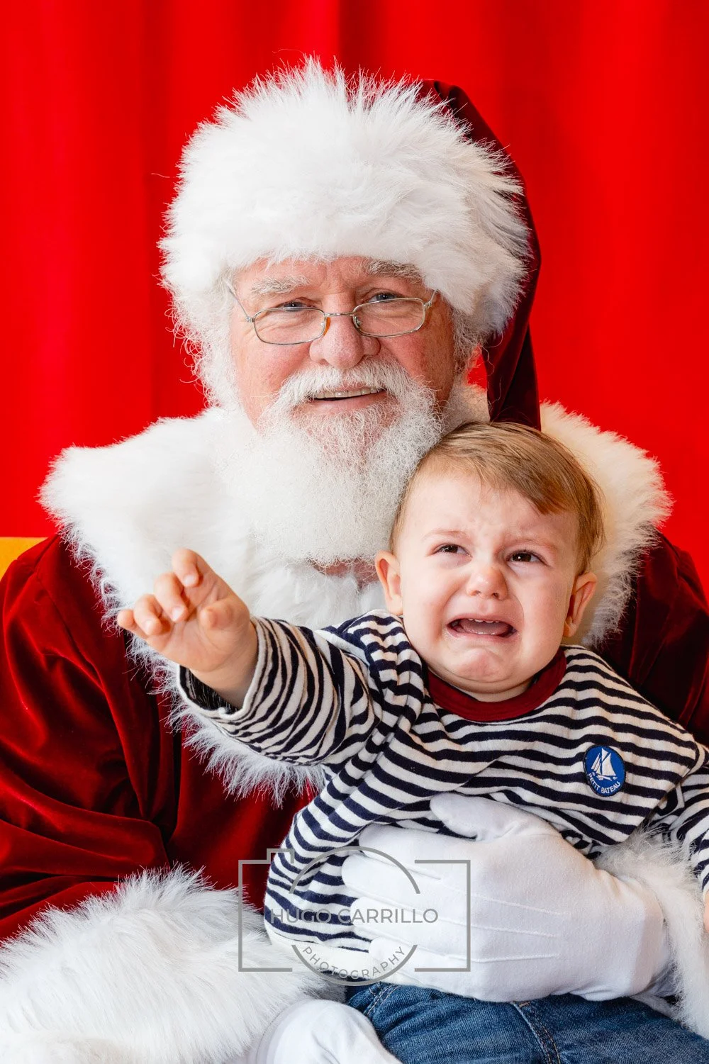 A man dressed as Santa Claus holding a crying young boy in front of a red background.