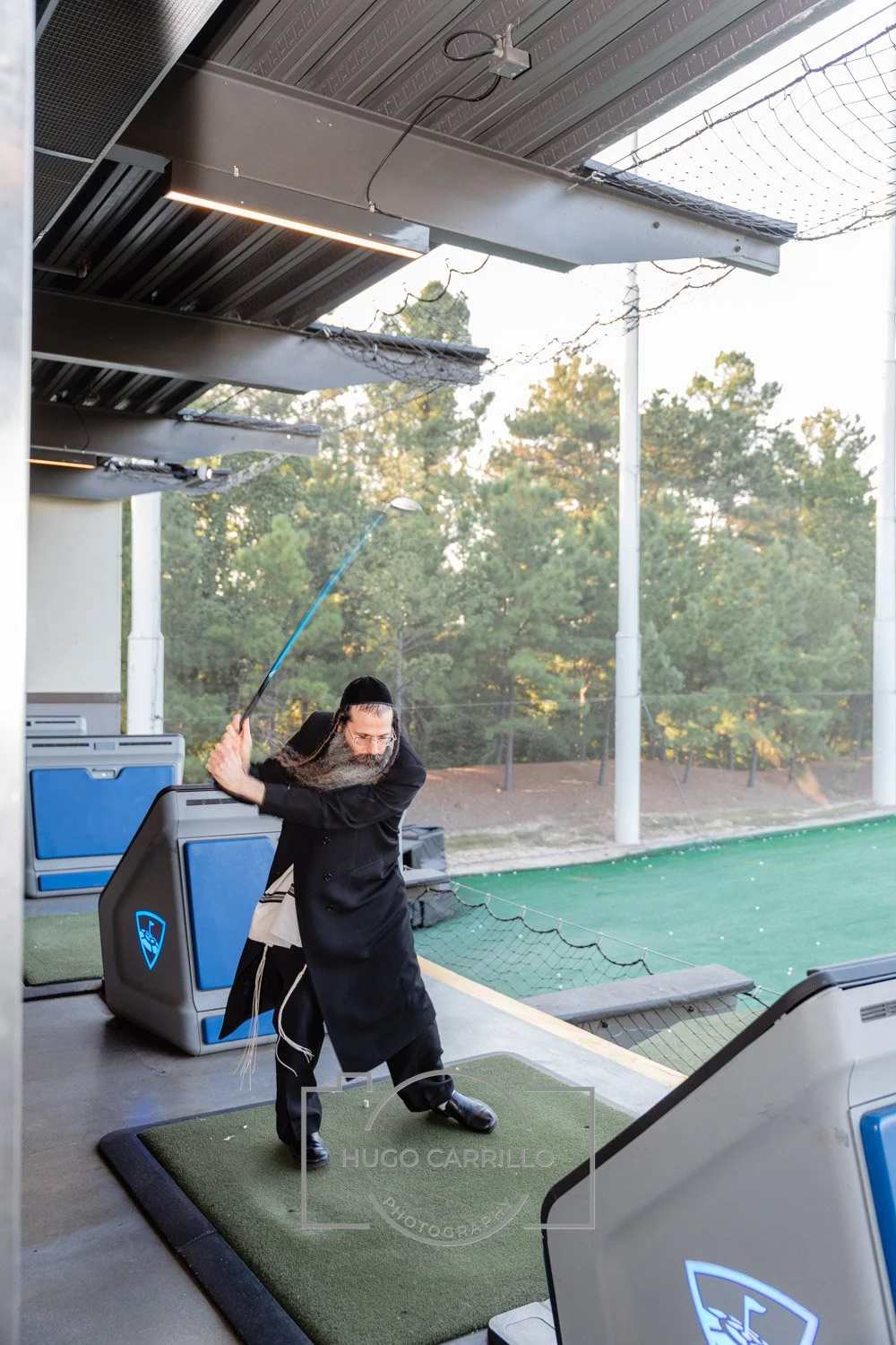 A man with a long beard and wearing traditional Jewish attire is practicing his golf swing at a driving range. He is standing on a mat, holding a golf club, and appears to be in mid-swing.