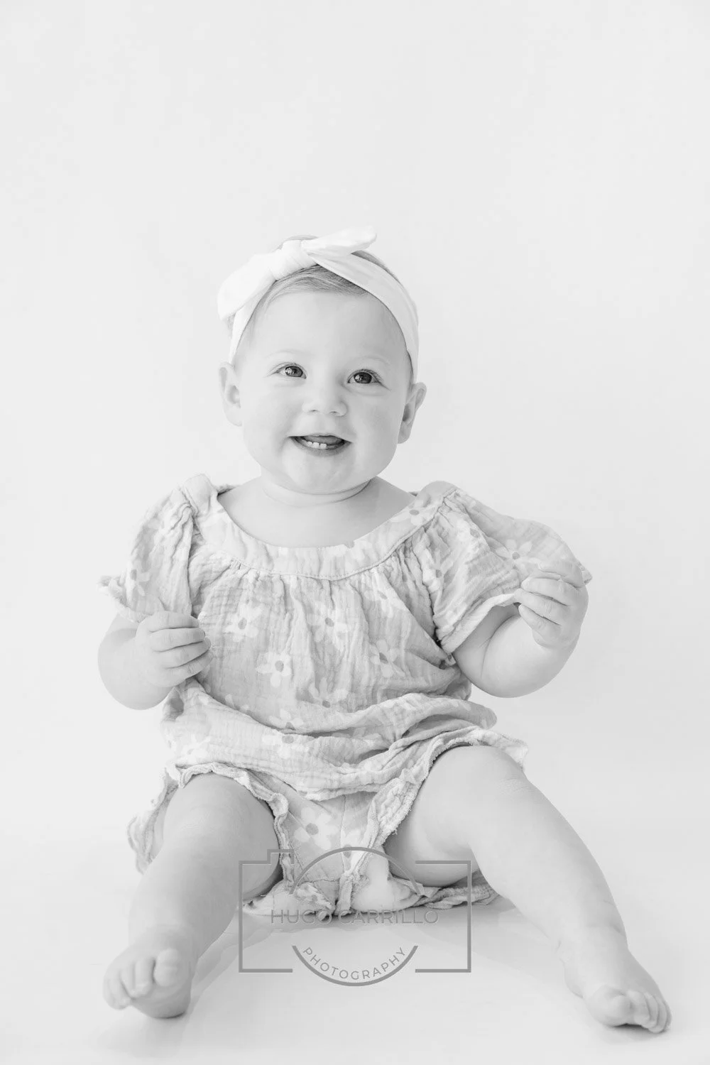 Black and white photo of a smiling baby girl sitting, wearing a headband with a bow and a patterned dress.
