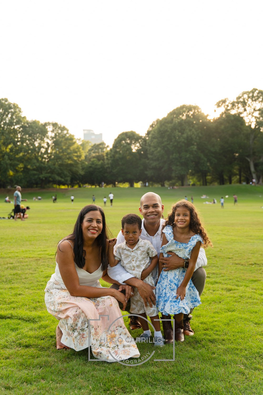 A family of five posing in a park with green grass and trees, some people in the background enjoying the outdoors, and a cityscape in the distance at sunset.
