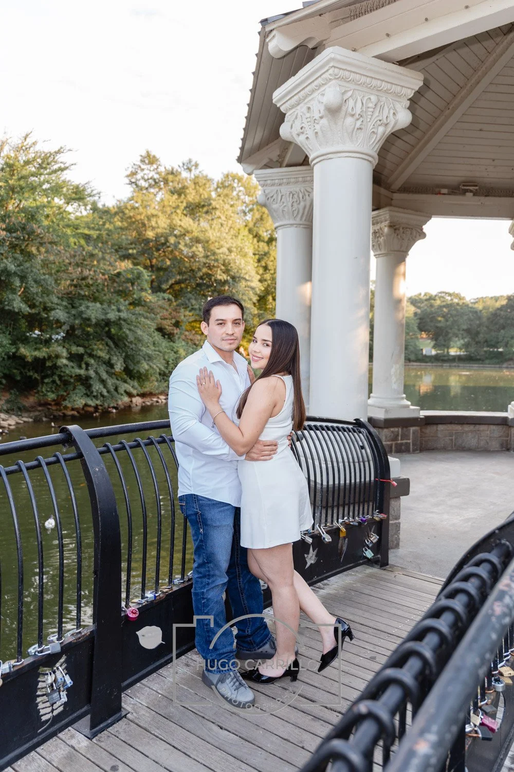 A couple is posing on a park bridge with lock love padlocks, a lake, trees, and a decorated pavilion in the background during the day.