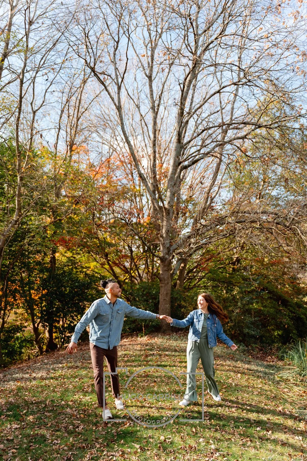 A man and woman in denim jackets hold hands and dance outdoors in a park with autumn foliage, leaf-covered ground, and a large, mostly bare tree.