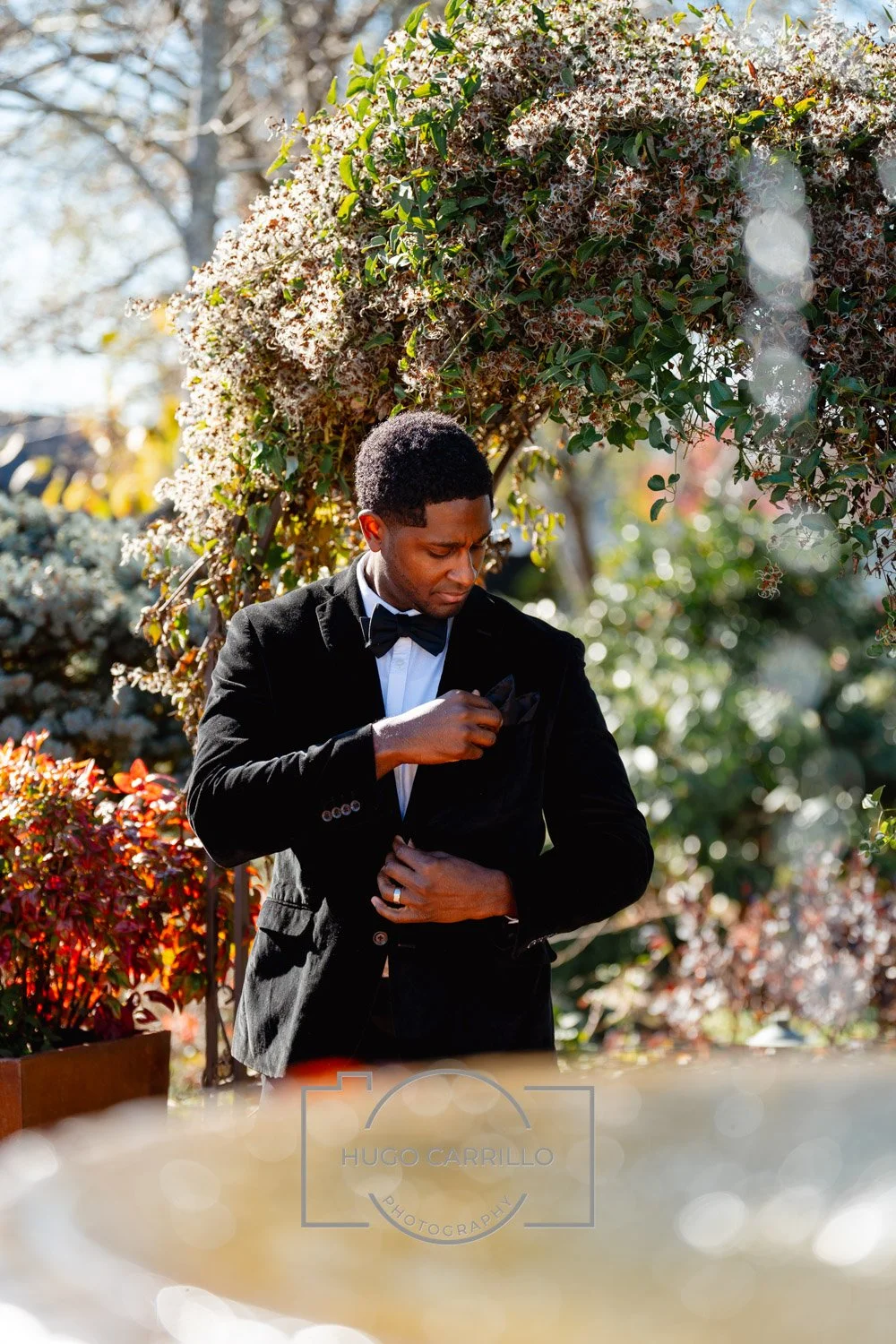 A man dressed in a black tuxedo with a bow tie adjusting his tuxedo jacket in an outdoor garden setting with a floral arch and autumn-colored plants.