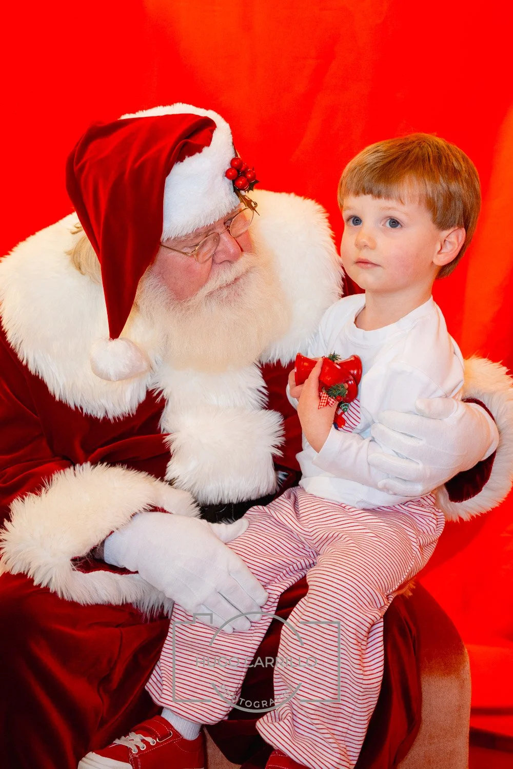 A young boy sitting on Santa Claus's lap holding a red ornament with a bow and a small Christmas tree decoration, with a red background.