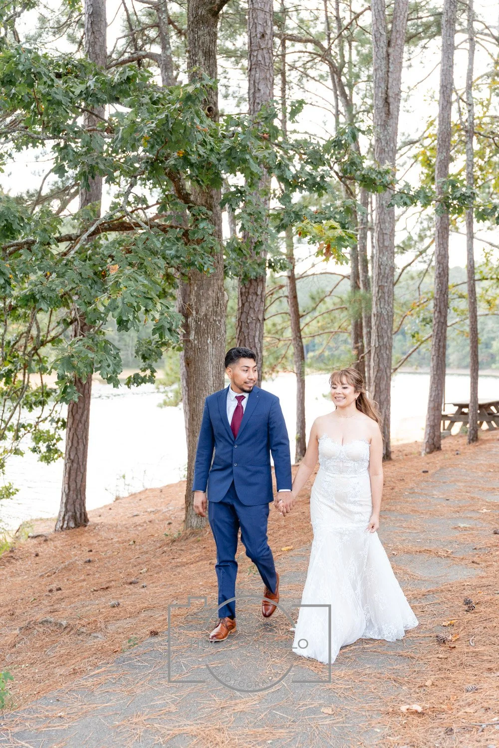 A newlywed couple holding hands and walking on a wooded path near a lake, surrounded by tall trees and picnic tables.