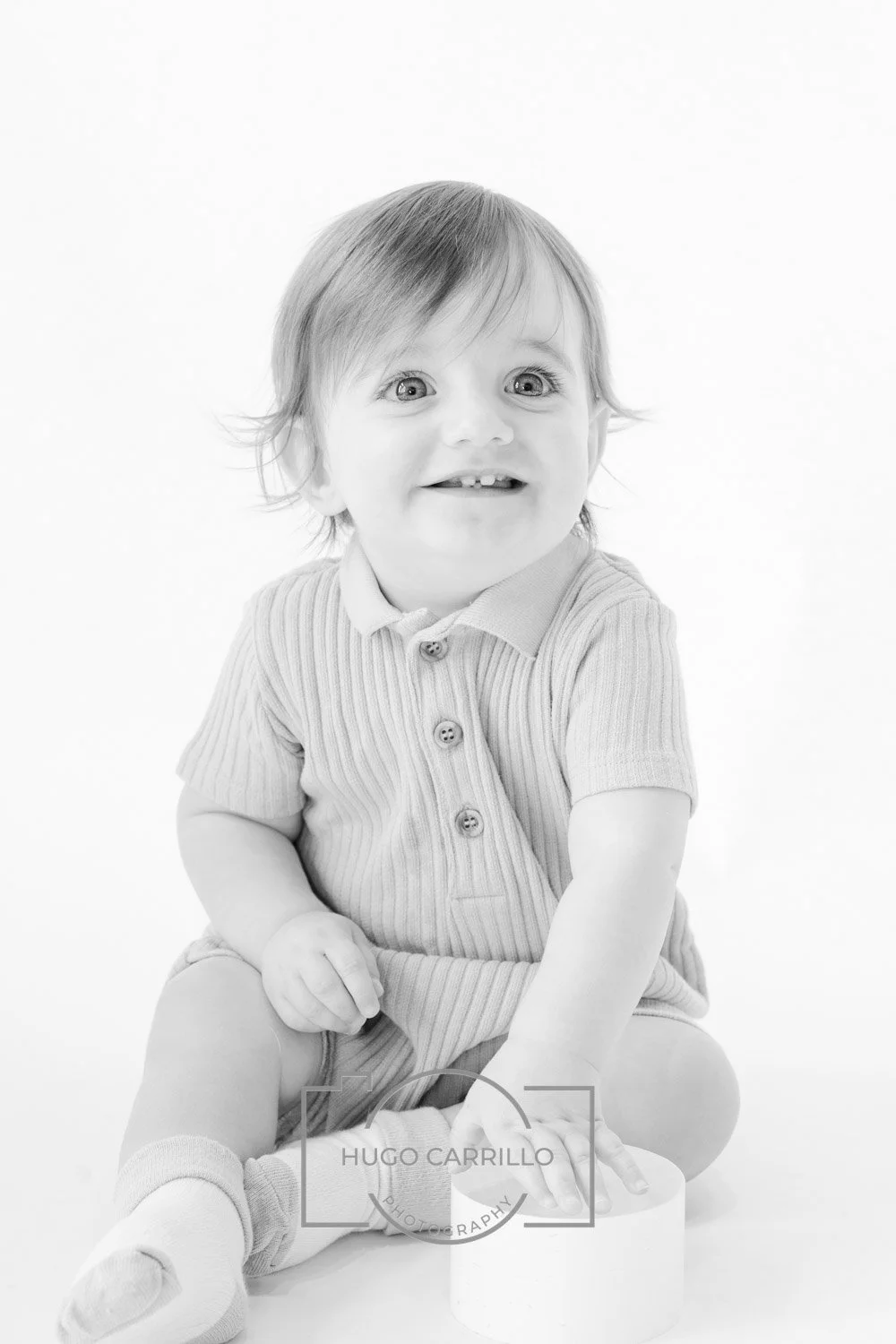 Black and white photo of a smiling toddler with light hair, wearing a short-sleeved, striped polo shirt and socks, sitting on the floor with legs crossed, touching a small cylindrical object, against a plain white background.
