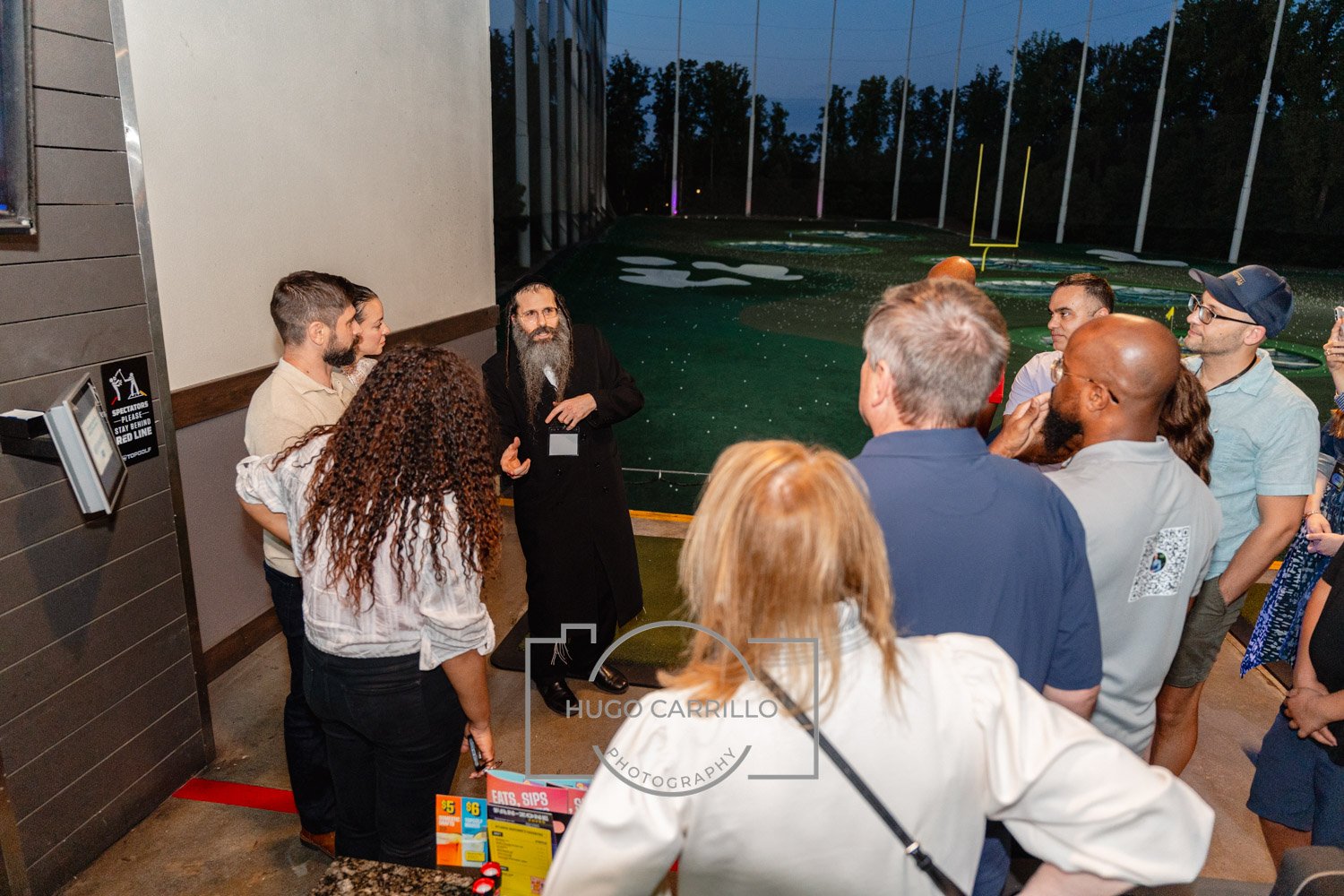 A group of people listening to a man with a beard and long hair who is speaking, in an indoor area near a large window or glass wall with an outdoor football field visible in the background.