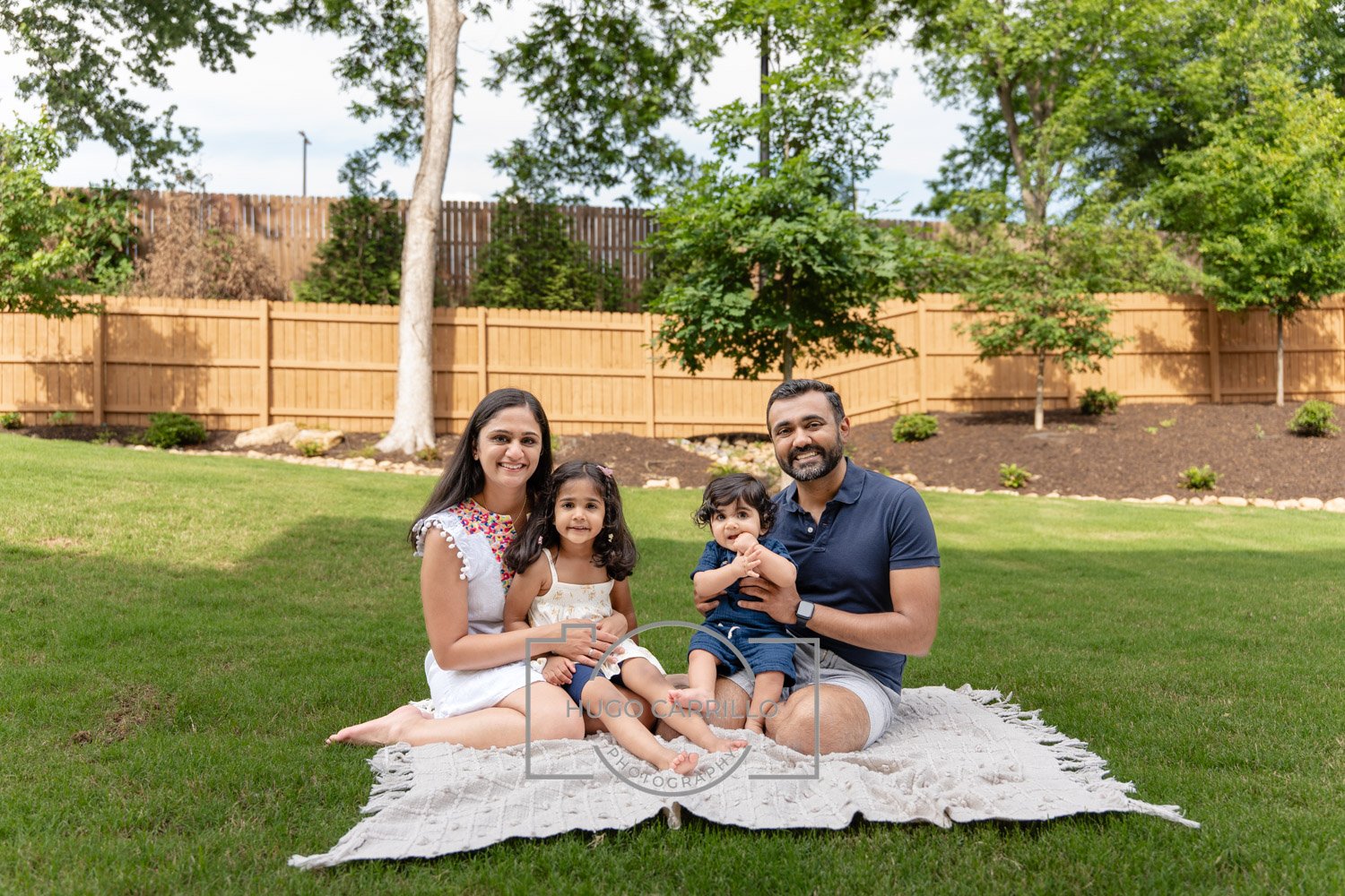 Family portrait of four sitting on a blanket in a backyard with green grass, trees, and a wooden fence, smiling at the camera.